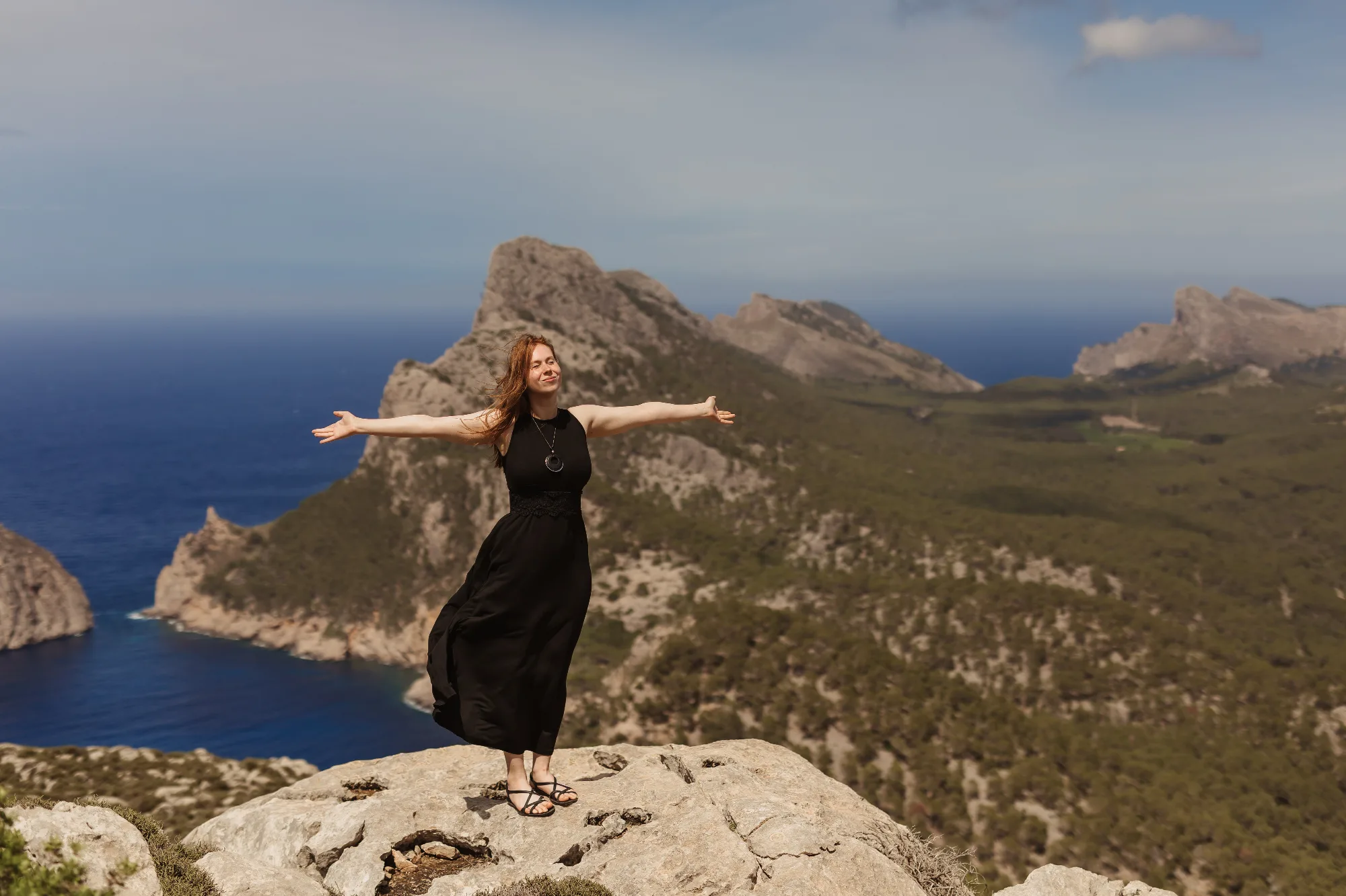 Young smiling woman with open arms and closed eyes in front of a beautiful mountain and sea landscape in Mallorca.